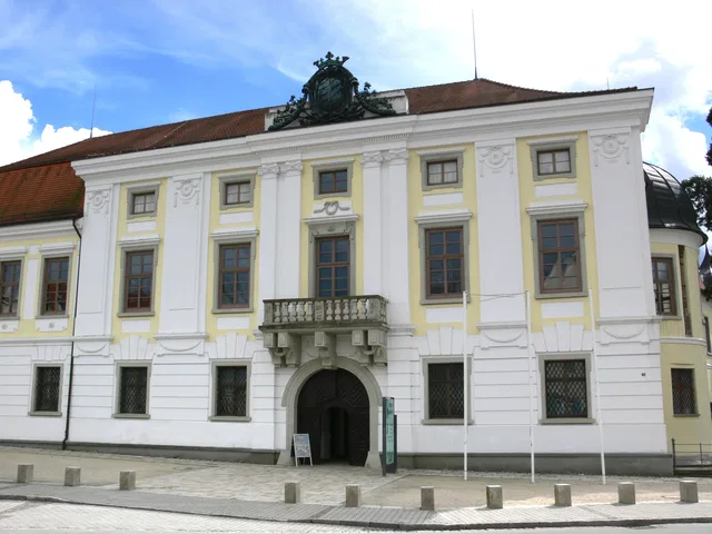 Historisches Gebäude mit gelber und weißer Fassade, zentraler Torbogen und Balkon