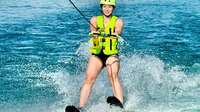 Person beim Wasserskifahren auf dem See, gelbe Schwimmweste und Helm, Spritzwasser um sie herum.