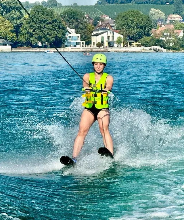 Person beim Wasserskifahren auf dem See, gelbe Schwimmweste und Helm, Spritzwasser um sie herum.