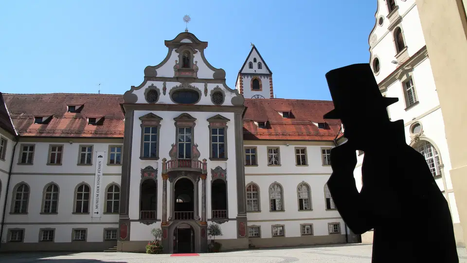 Füssen Klosterhof mit barocker Fassade, Innenhof und einer silhouettierten Figur im Vordergrund.