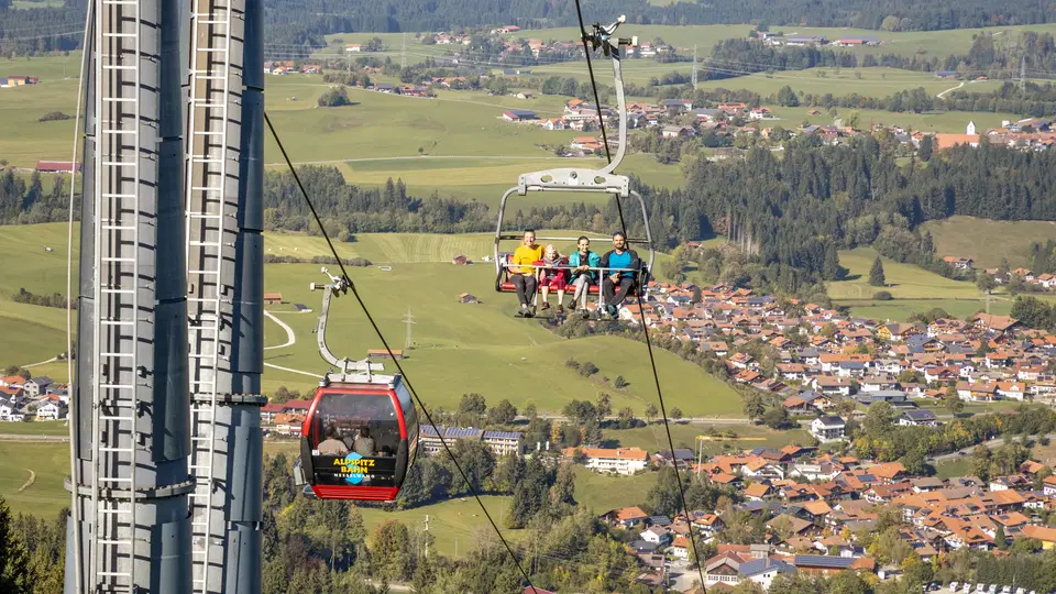 Personen in einer Seilbahnkabine über einer ländlichen Umgebung mit Feldern und Dorf im Hintergrund.