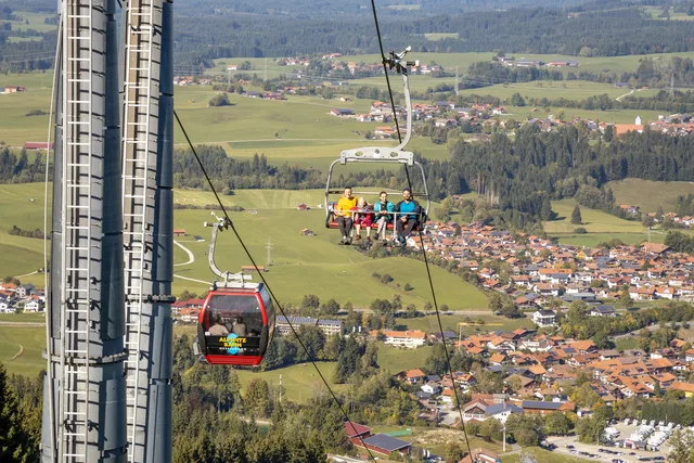 Personen in einer Seilbahnkabine über einer ländlichen Umgebung mit Feldern und Dorf im Hintergrund.