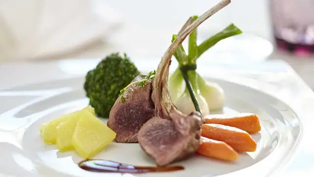 Lammkotelett mit Gemüse (Brokkoli, Karotten), Ananaswürfel und Soße auf weißem Teller.