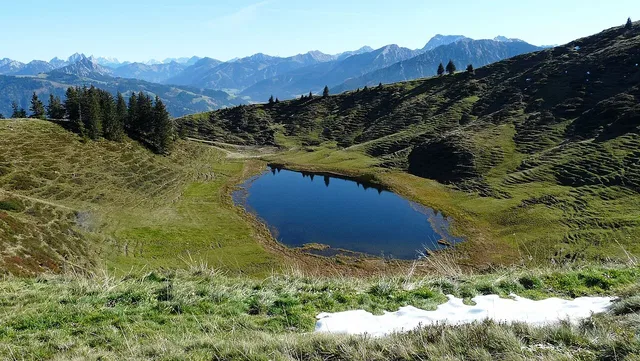 Kleiner Bergsee in einer grünen Hügellandschaft mit Baumgruppen vor einer Bergkette.