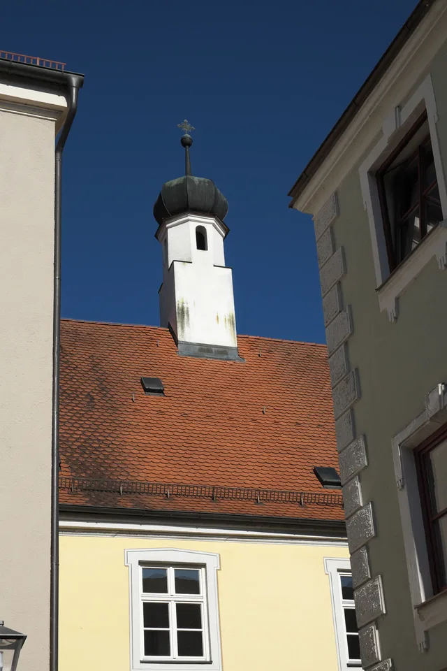 Ansicht eines Gebäudes mit rotem Ziegeldach und kleinem Turmaufsatz zwischen zwei Häusern.