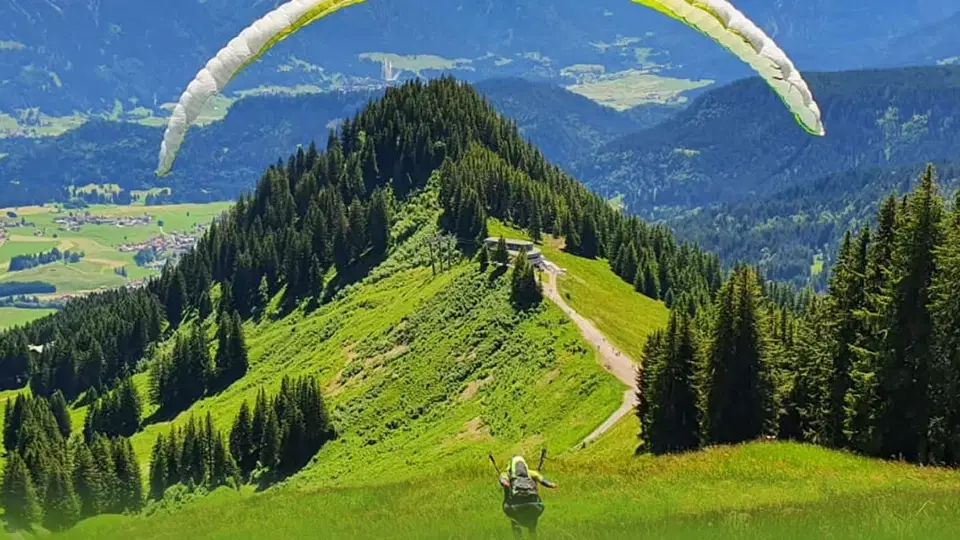 Paragleiter startet von einer grünen Bergwiese mit Wald und Bergpanorama.