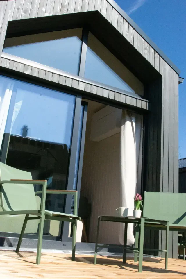 Moderne Holzhütte mit großen Glasfenstern und Terrasse mit grünen Gartenstühlen.