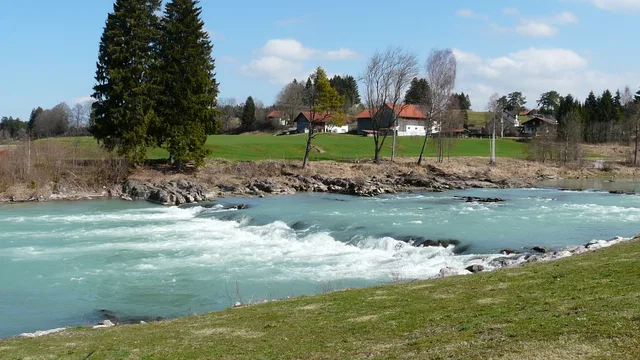 Flussufer mit türkisfarbenem Wasser, Bäumen und Häusern im Hintergrund.