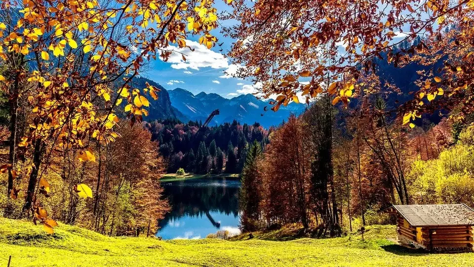 Herbstliche Landschaft mit See, Wald und Bergkette; rechts eine kleine Holzhütte.