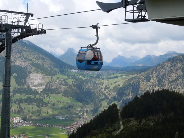 Symbolbild: Blaue Seilbahn-Gondel fährt über Berglandschaft mit Tal. Keine Originalaufnahme dieses Eintrags.