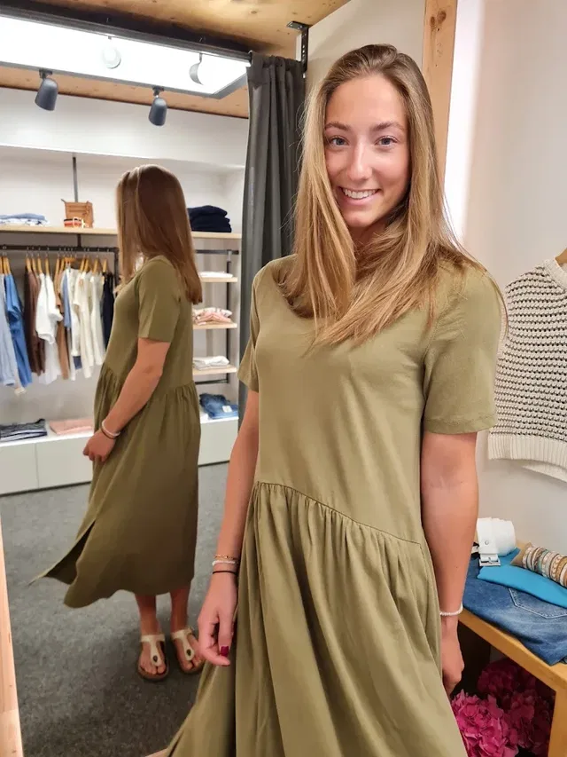 Junge Frau in olivegrünem Kleid lächelt im Kleidergeschäft; Spiegel zeigt ihre Rückenansicht.