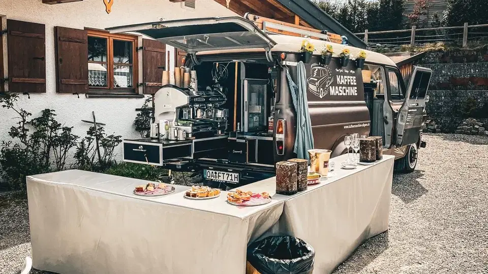 Kaffee-Mobil mit ausgestatteter Bar und Kuchenbuffet vor einem Haus.