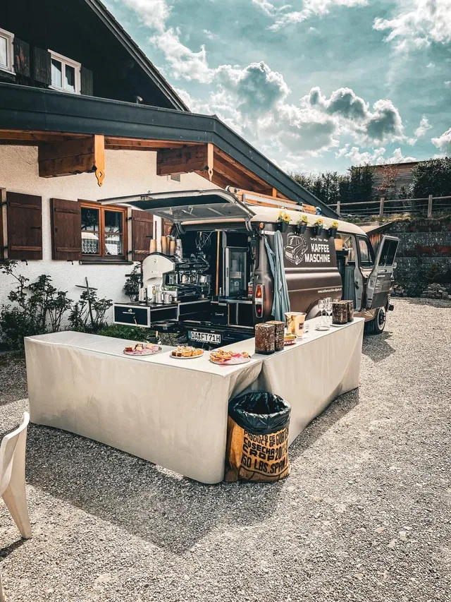 Kaffee-Mobil mit ausgestatteter Bar und Kuchenbuffet vor einem Haus.