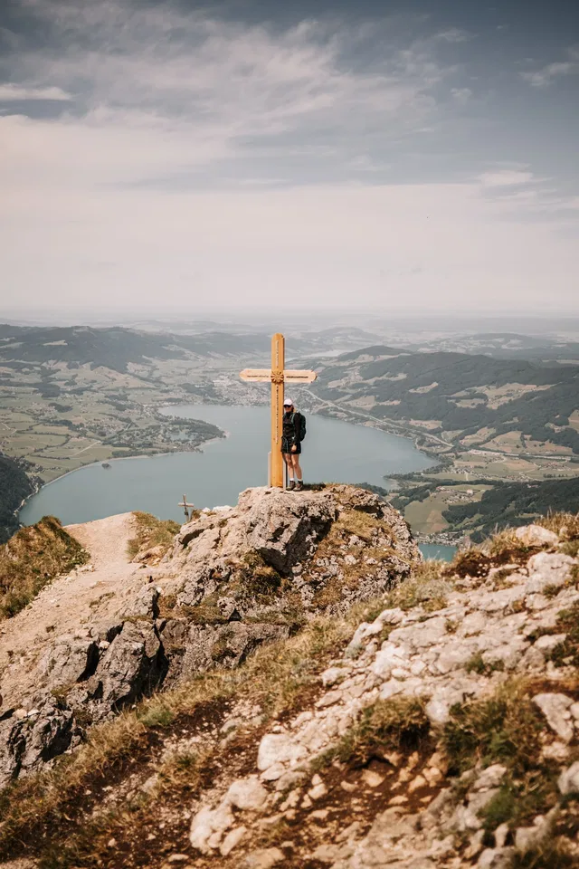 Person steht an Felsenkante neben Holzkreuz mit Blick auf See und Berge.