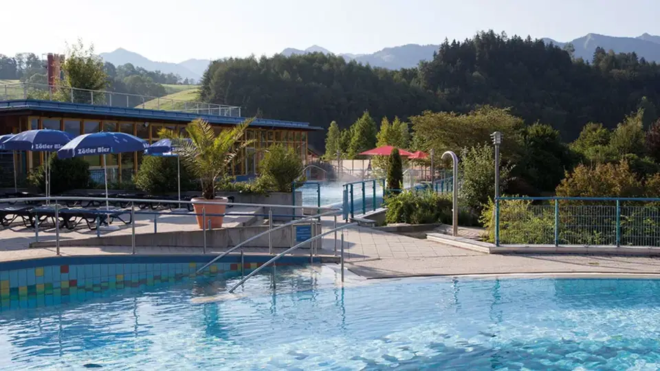 Außenszene eines Freibads mit Becken, Liegen, Sonnenschirmen und Terrassenbereich, umgeben von Bämen