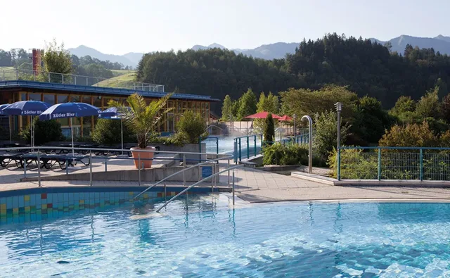 Außenszene eines Freibads mit Becken, Liegen, Sonnenschirmen und Terrassenbereich, umgeben von Bämen