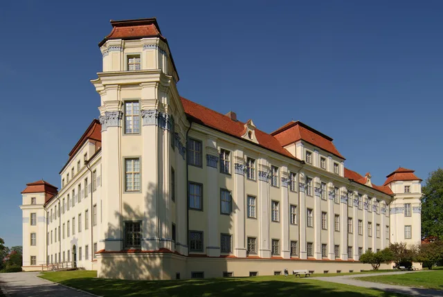 Großes Schloss mit gelber Fassade, roten Dächern und Türmen vor blauem Himmel.