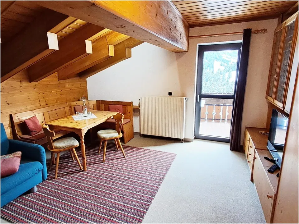 Dachgeschoss-Wohnzimmer mit Holzbalkendecke, Esstisch und Stühle, blaues Sofa, Teppich und Balkontür