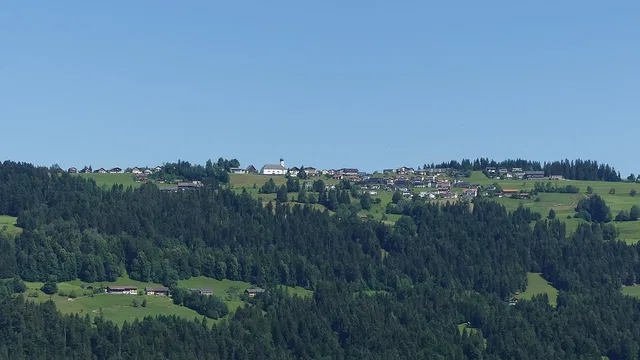 Landschaft mit grünen Hügeln, Waldflächen und verstreuten Häusern am Horizont.