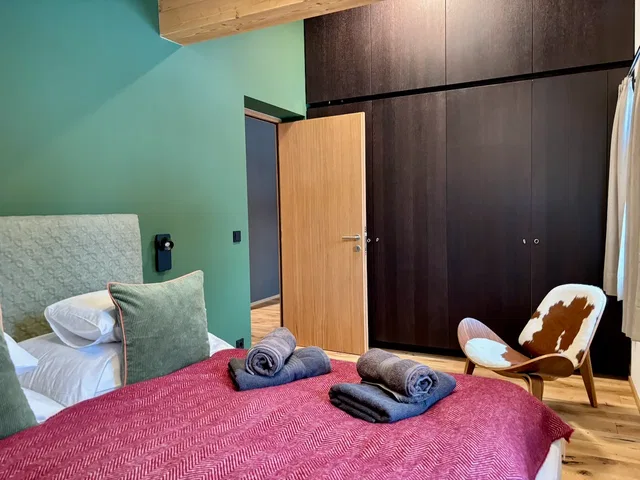 Schlafzimmer mit grüner Wand, rotem Bettüberwurf und dunklem Kleiderschrank, offener Tür und Lounge‑