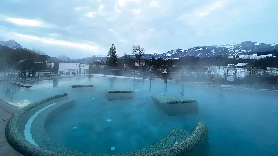 Außenbecken eines Thermalbads mit dampfender Oberfläche und Blick auf schneebedeckte Berge.