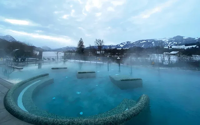 Außenbecken eines Thermalbads mit dampfender Oberfläche und Blick auf schneebedeckte Berge.