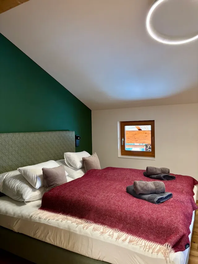 Schlafzimmer mit grünem Wandakzent hinter dem Bett, roter Decke, weißen Kissen und zwei auf dem Bett