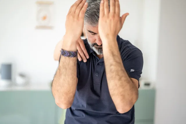 Mann mit grauem Bart greift sich den Kopf, wirkt gestresst in hellem Innenraum.