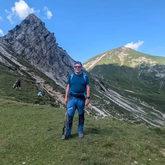 Mann in blauem Wanderoutfit mit Stock steht auf grasigem Bergpfad vor felsigen Gipfeln.