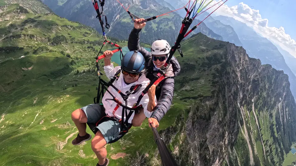 Gast in hellem Shirt mit Pilot beim Tandemflug vor einer felsigen Bergwand.
