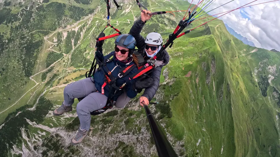 Gast mit blauer Helmhaube und Pilot beim Tandemflug über grüne Hänge.