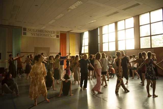 Eine große Gruppe Tänzerinnen und Tänzer erlebt Ecstatic Dance in einer hellen Sporthalle mit bunten Vorhängen und Banner im Hintergrund.