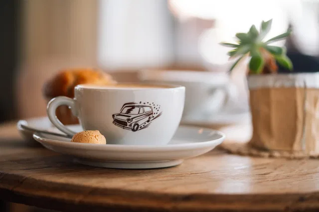 Weiße Kaffeetasse mit Autoskizze auf Untertasse, daneben Keks, Holztisch, Pflanztopf im Hintergrund