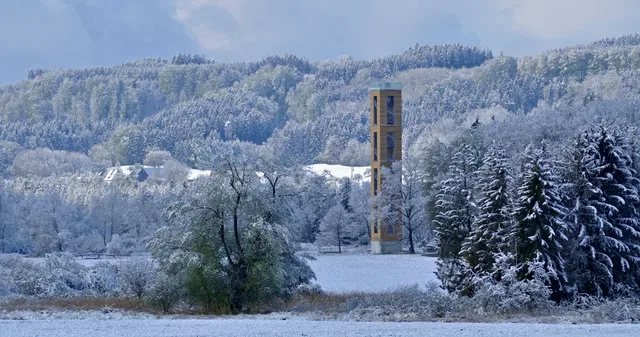 Winterliche Landschaft mit Schnee, Bäumen und einem gelb-braunen Turm im Zentrum.