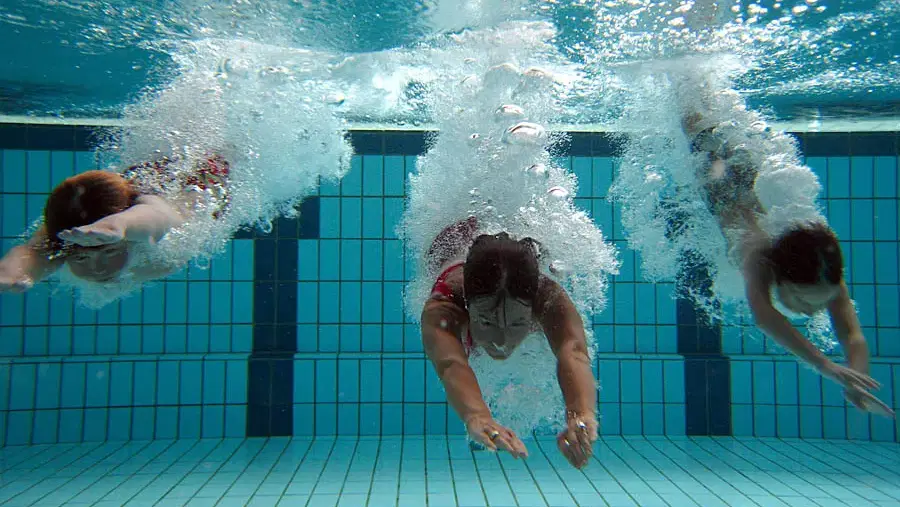 Drei Schwimmer tauchen im Beckenboden eines Pools, Luftblasen steigen auf.