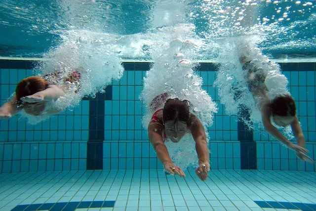 Drei Schwimmer tauchen im Beckenboden eines Pools, Luftblasen steigen auf.
