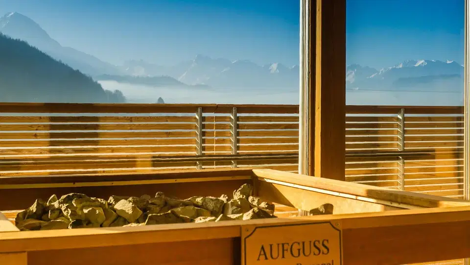 Sauna mit Holzofen und Steinen; Blick auf Berge durch Glasfenster.