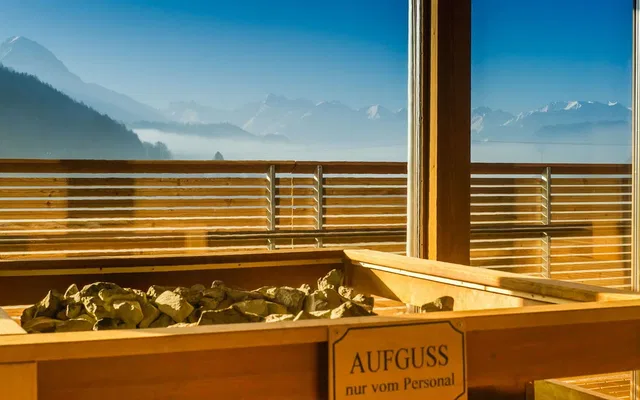 Sauna mit Holzofen und Steinen; Blick auf Berge durch Glasfenster.