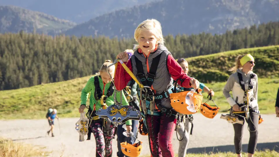Kinder mit Kletterausrüstung und Helmen gehen bergauf in einer Berglandschaft.