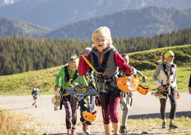 Kinder mit Kletterausrüstung und Helmen gehen bergauf in einer Berglandschaft.