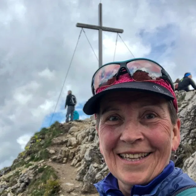 Porträt einer lächelnden Frau auf einem felsigen Berggipfel mit Kreuz im Hintergrund.