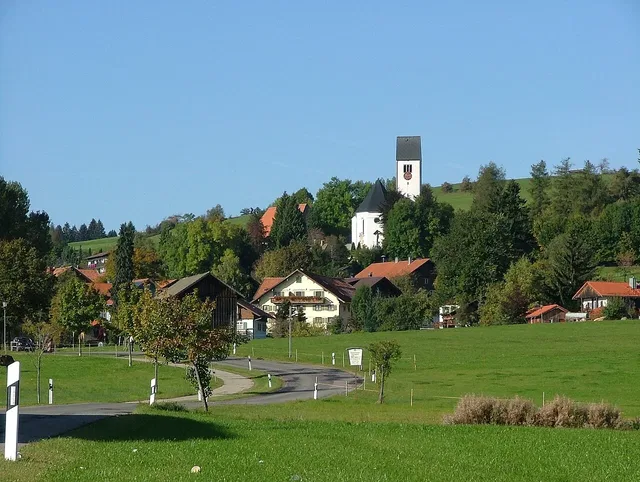 Kleines Dorf mit Kirchturm auf einem Hügel, umgeben von Grünflächen und Bäumen.