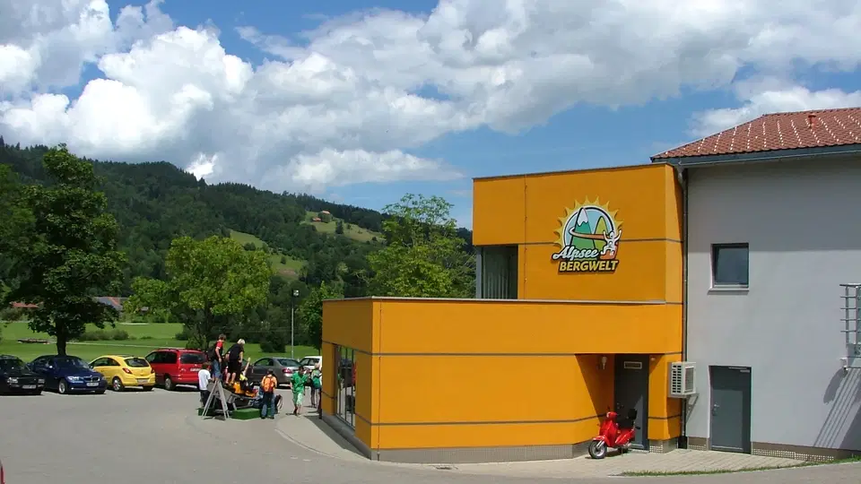 Gelb-orange Gebäude mit Bergwelt-Logo am Parkplatz, umliegende Berglandschaft und Autos.