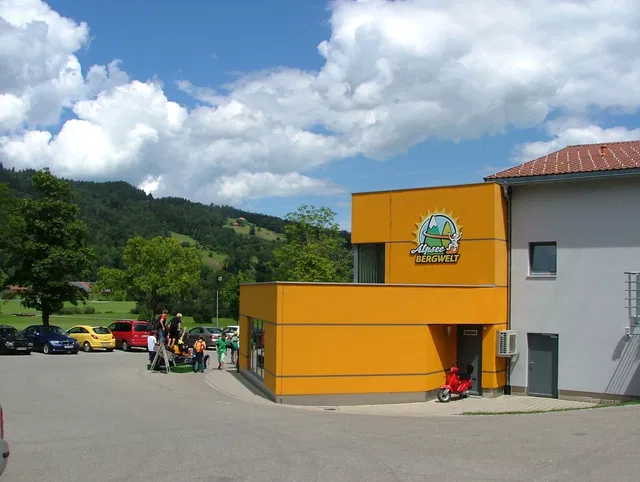 Gelb-orange Gebäude mit Bergwelt-Logo am Parkplatz, umliegende Berglandschaft und Autos.