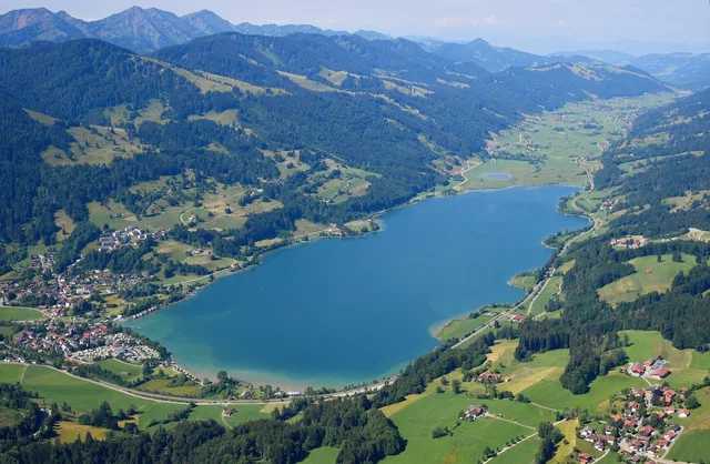 Luftaufnahme eines türkis-blauen Sees im Alpengebiet mit umliegenden Dörfern und grünen Wiesen.