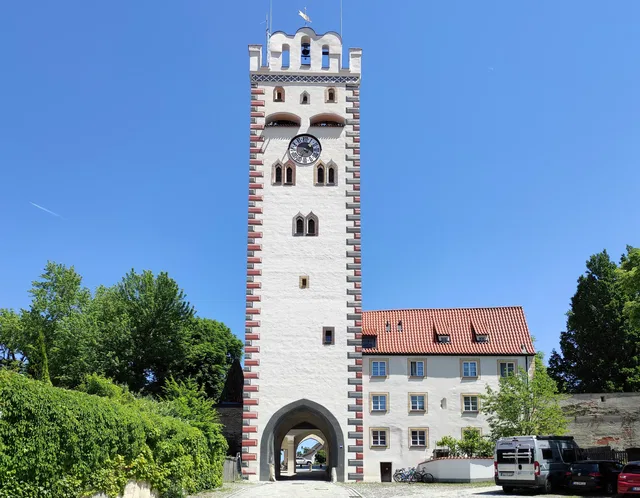 Weißer Turm mit roten Zierleisten, Torbogen am Grund, Uhr am Turm, daneben Gebäude und Bäume