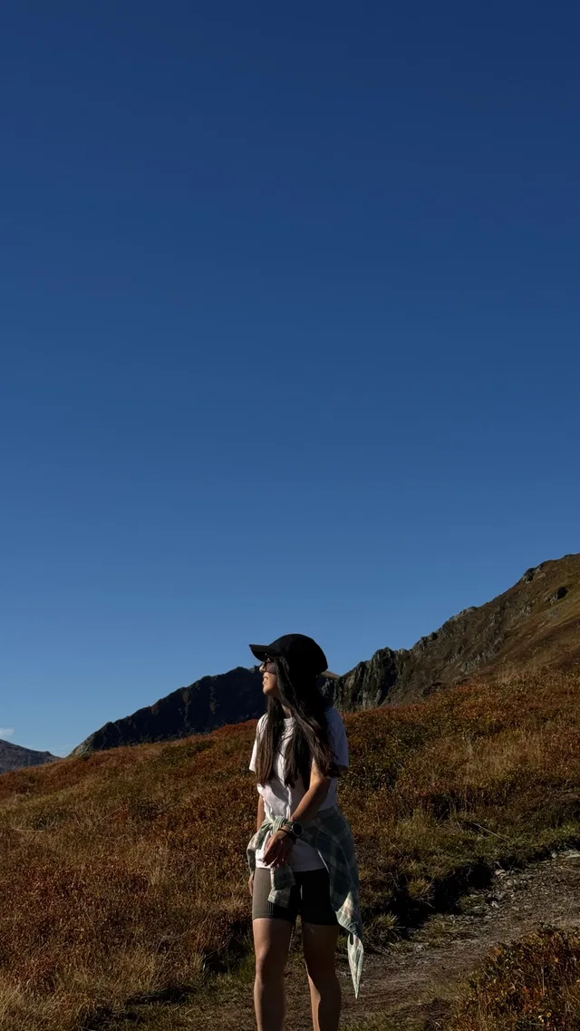 Person mit Hut am Wanderweg in bergiger Herbstlandschaft unter blauem Himmel.