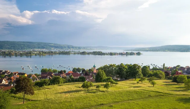 Landschaft mit Dorf am Seeufer, grünen Feldern und Booten auf dem Wasser.