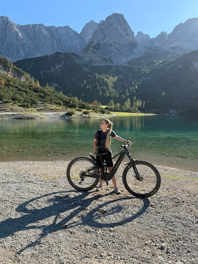 Frau mit E-Bike am Ufer eines Sees vor Bergen