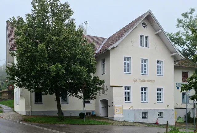 Mehrstöckiges, helles Haus mit rotem Ziegeldach und Großbaum davor.